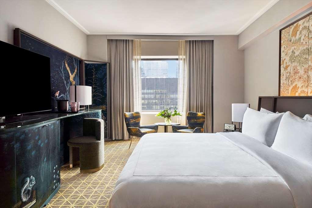 Hotel Park Lane New York Camera foto The photo shows a stylish hotel room featuring a large bed with white bedding. In the background, there is a large window with curtains that lets in natural light. A small table with two chairs is positioned near the window, suggesting a cozy seating