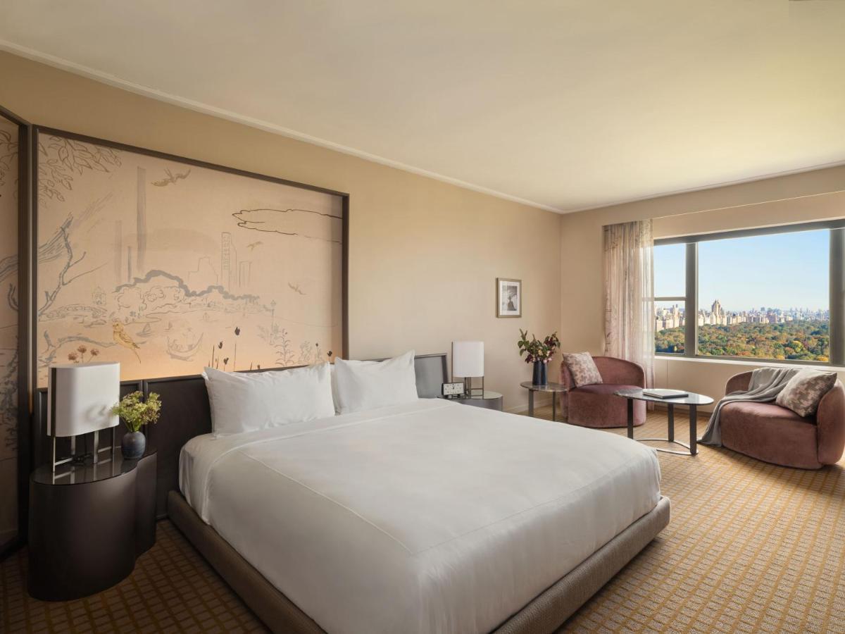 Hotel Park Lane New York Esterno foto The photo shows a modern hotel room featuring a large bed with white linens placed on a stylish platform base. Behind the bed, there is an artistic wall mural, and the decor has a neutral color palette. To the side, there are two comfortable chairs w