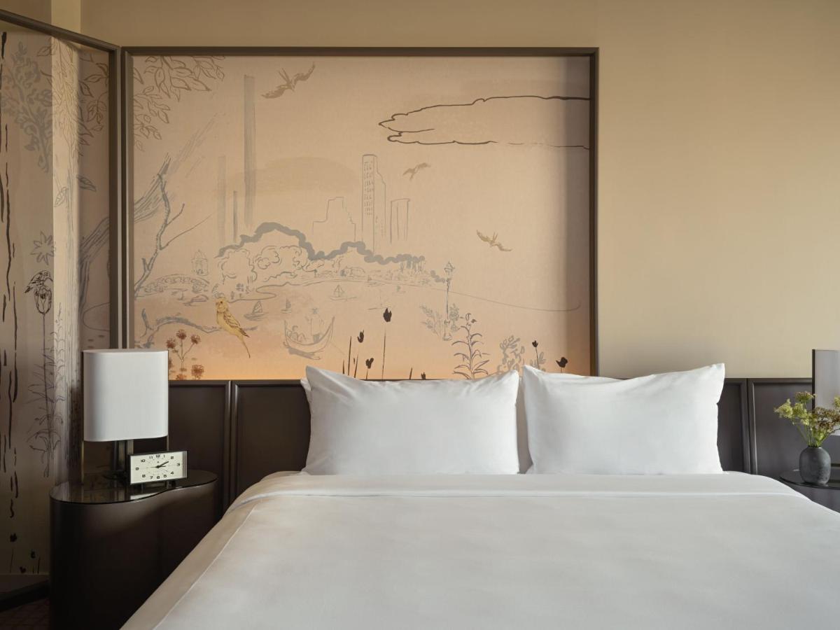 Hotel Park Lane New York Esterno foto The photo depicts a modern hotel room featuring a plush bed with white linens and two pillows. Behind the bed is an artistic wall mural that appears to illustrate a serene landscape with subtle outlines of trees and clouds. On either side of the bed,