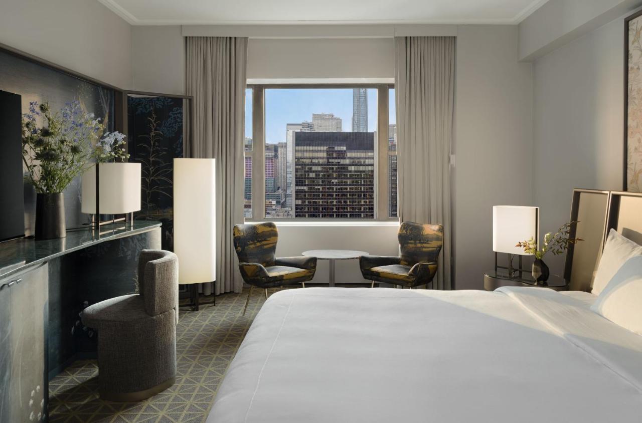 Hotel Park Lane New York Esterno foto The photo shows a stylish hotel room featuring a large bed with white linens. There are two modern armchairs beside a small round table, positioned near a window that offers a view of the cityscape. Natural light flows into the room, highlighting the