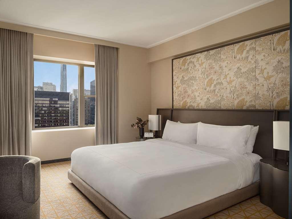 Hotel Park Lane New York Camera foto The photo shows a modern hotel room featuring a large bed with white bedding against a decorative headboard. There are two bedside lamps on either side of the bed. The room has light-colored walls and soft carpeting. A large window allows natural lig