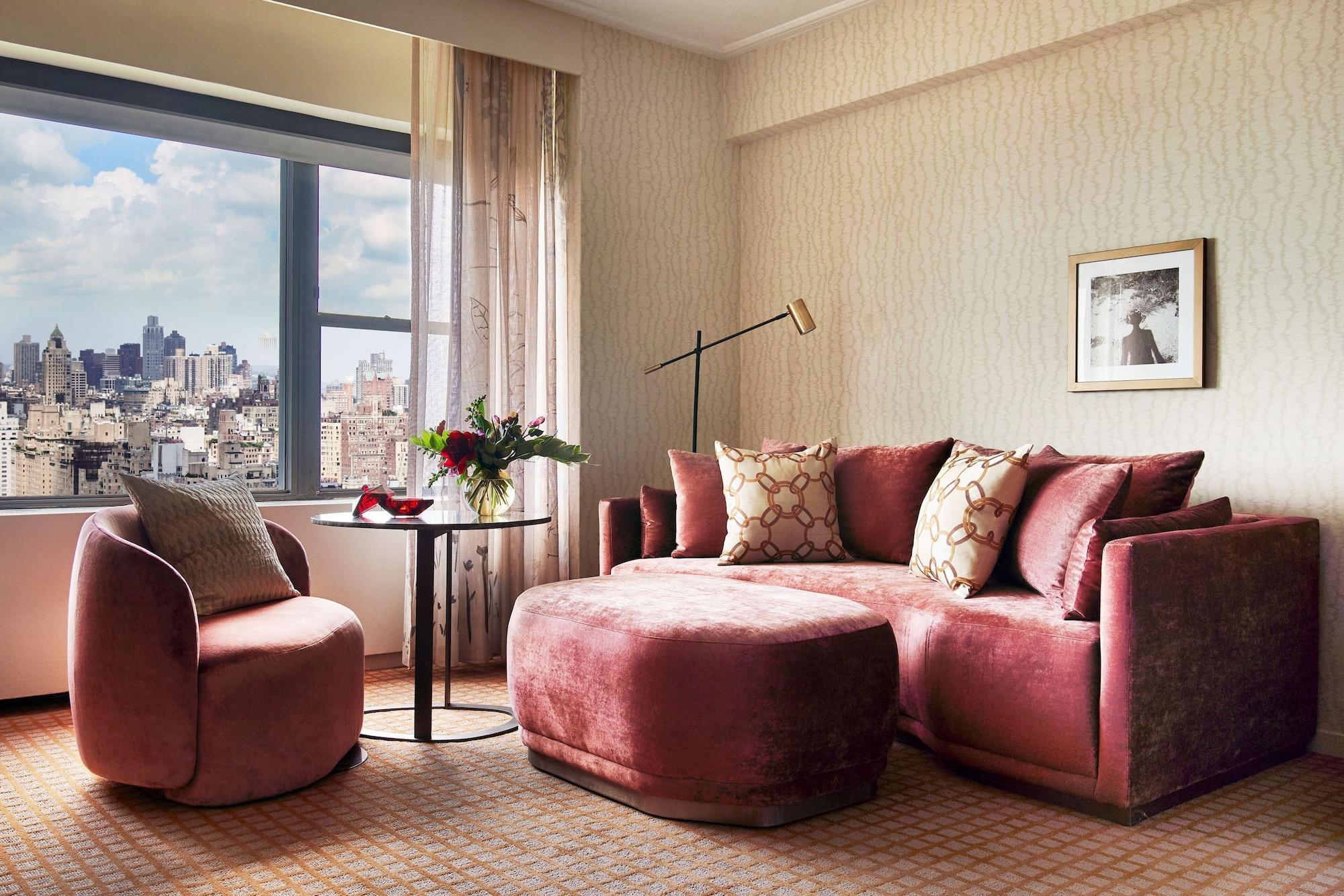 Hotel Park Lane New York Esterno foto The photo shows a modern indoor living space, likely a hotel room or a stylish apartment. There is a plush, curved sectional sofa in a soft pink hue, adorned with decorative throw pillows. A round table sits in front of the sofa, accompanied by a sin