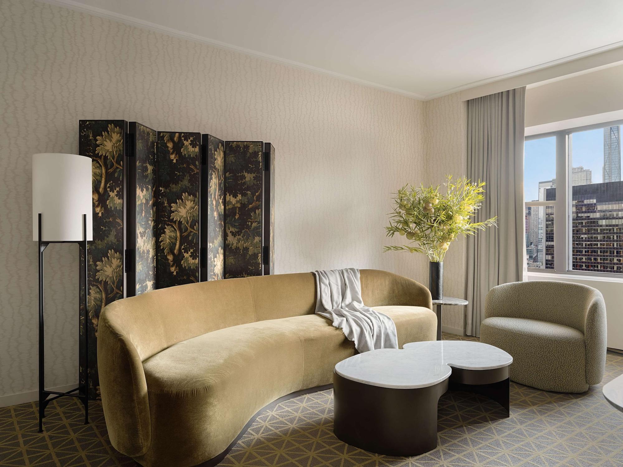 Hotel Park Lane New York Esterno foto The photo shows a stylish interior space that appears to be a living area or lounge. There is a large, curved beige sofa with a throw blanket draped over it. In front of the sofa are two coffee tables, one round and white and the other oval and dark.