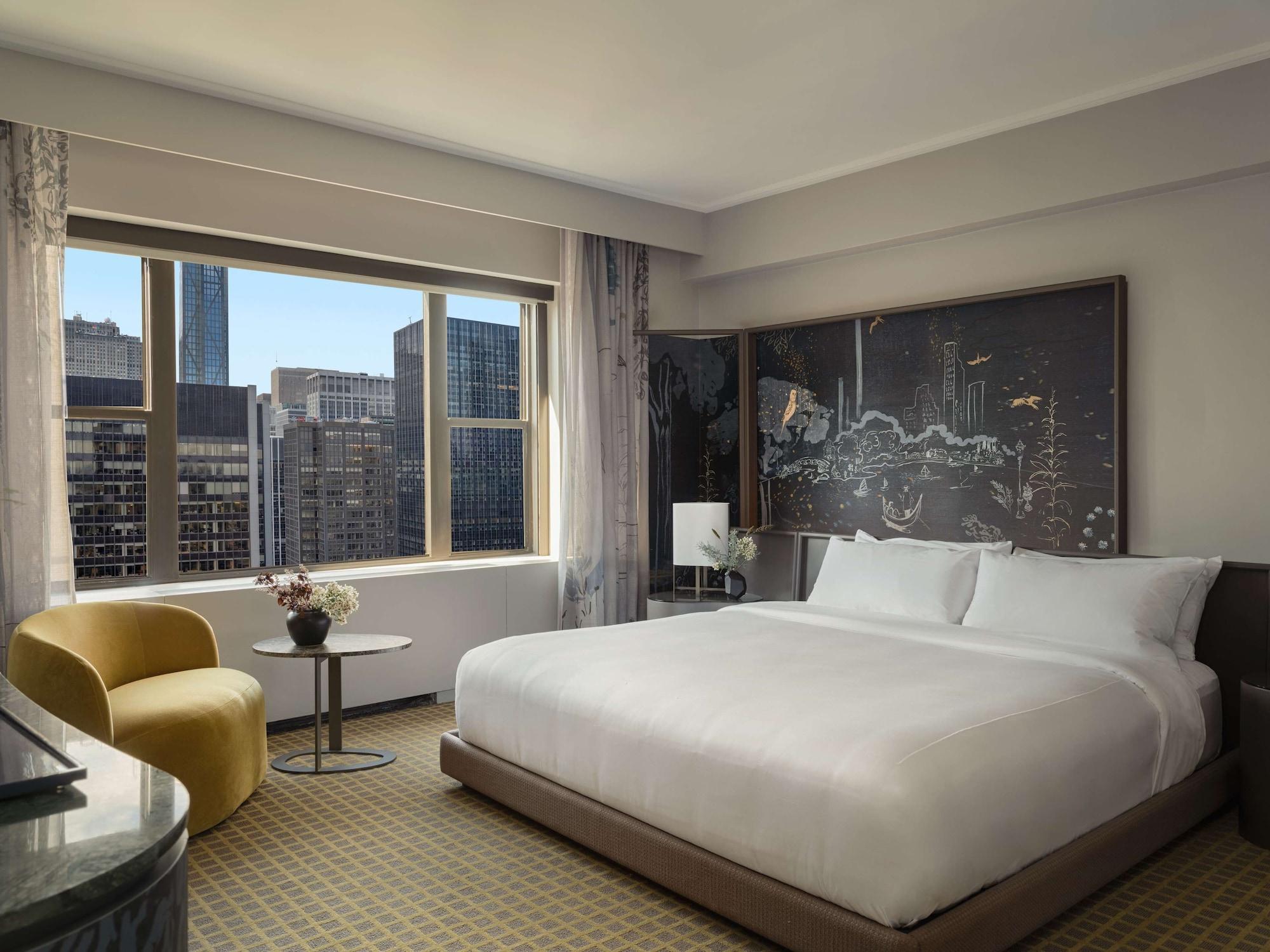 Hotel Park Lane New York Esterno foto The photo shows a modern hotel room featuring a large bed with white linens, a stylish yellow chair, and a small side table. There is a large window with a view of a cityscape, allowing natural light to fill the room. The decor includes a piece of ar