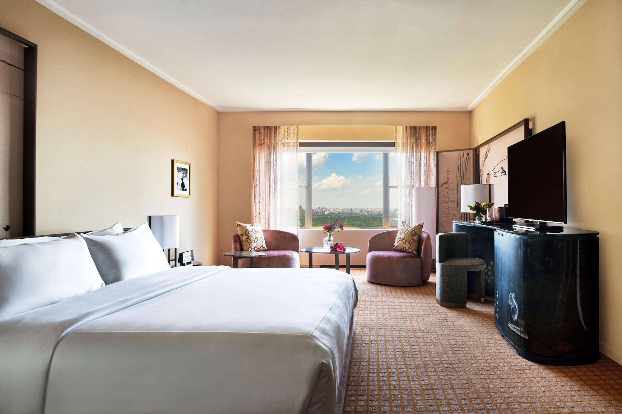 Hotel Park Lane New York Esterno foto The photo shows a spacious hotel room with a warm beige color scheme. There is a large king-sized bed with white linens prominently placed in the foreground. To the side, there are two comfortable-looking chairs facing a small table. 

Across from th