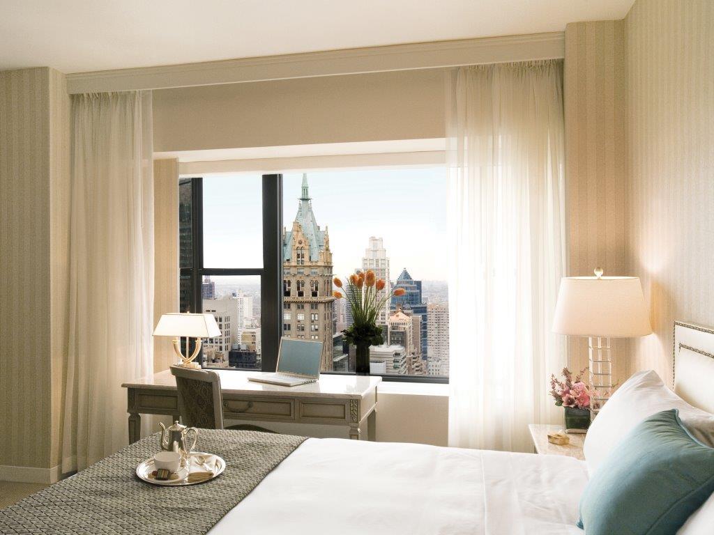 Hotel Park Lane New York Esterno foto The photo depicts a stylish hotel room with a neatly made bed featuring white linens and a decorative throw. There's a modern desk near a large window, which offers a view of a city skyline with notable buildings. The room features soft color tones a
