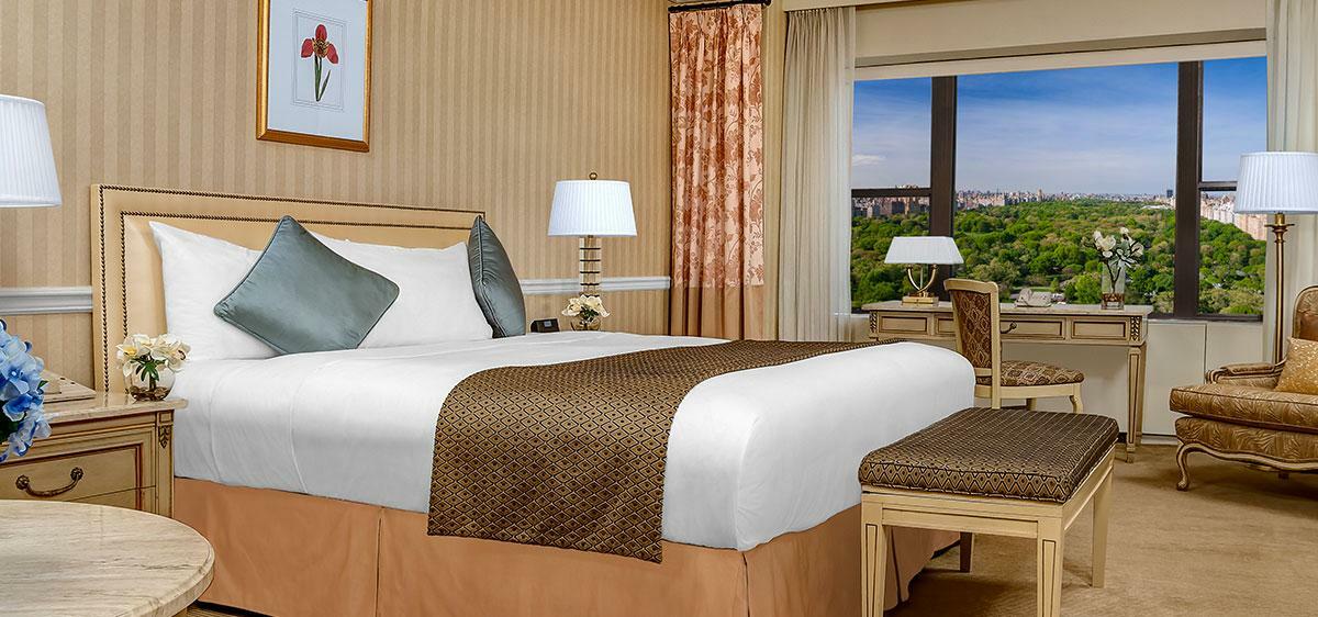 Hotel Park Lane New York Esterno foto The photo shows a well-furnished hotel room. It features a large bed with a white comforter and decorative pillows. There's a small stool at the foot of the bed, and a bedside lamp on a nightstand. The room has warm tones with striped wallpaper and a