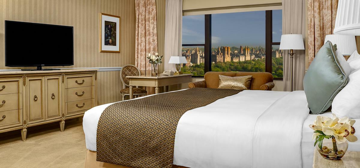 Hotel Park Lane New York Esterno foto The photo shows an elegantly designed hotel room. In the foreground, there is a large bed with a white comforter and a decorative brown throw blanket. Next to the bed, there is a comfortable-looking couch. A wooden nightstand and a lamp are visible. 