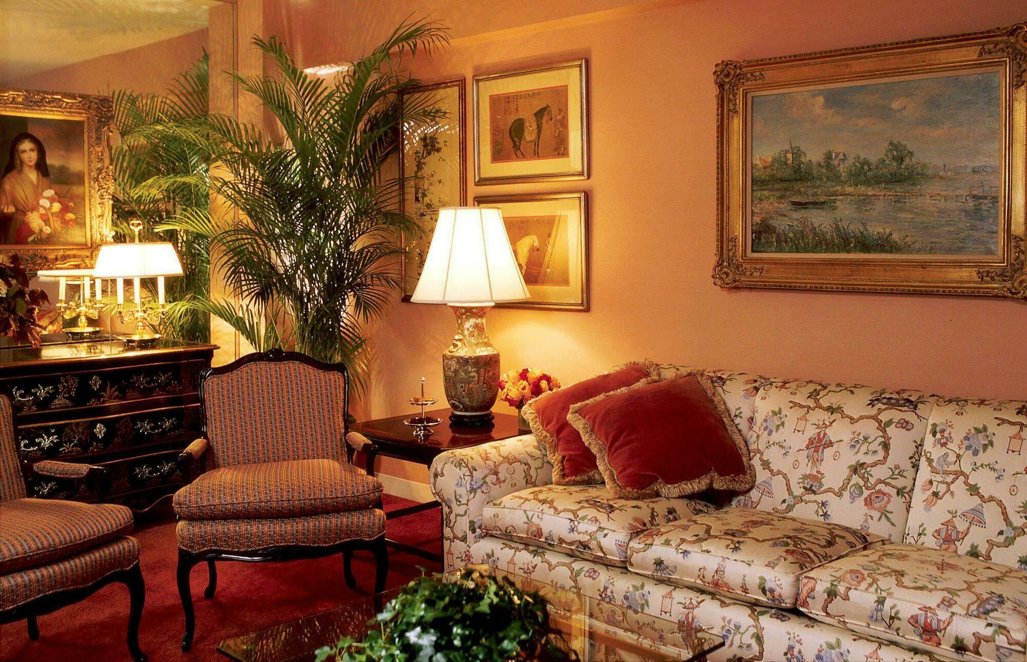 Hotel Park Lane New York Camera foto The photo shows a cozy living room setting. The walls are painted a warm peach color, and there are several framed paintings hanging on the walls. A floral-patterned sofa with plush cushions is positioned in the room, along with elegant armchairs tha